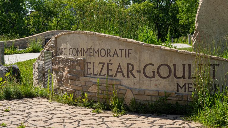 Rock and masonry sculpture shaped like a curved wall emblazoned with with large text reading “Parc commémoratif Elzéar Goulet.”