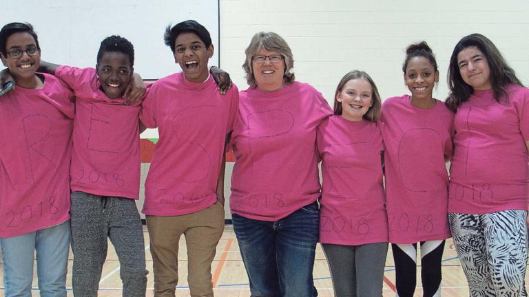 Six teenagers and a middle-aged woman stand with their arms around each other and are smiling for the camera. They are all wearing pink shirts. Partially obscured.