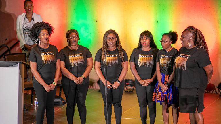 A group of six black performers standing on stage with colourful backlighting. Partially obscured.