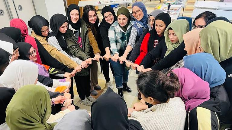 A group of smiling Muslim women in a circle with linked hands. Partially obscured.