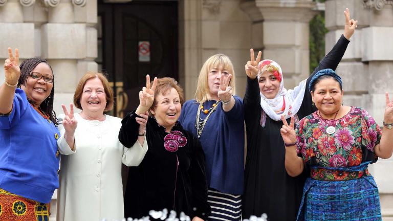 Six women are standing in front of what appears to be the entrance of a large stone building. They are all smiling and holding up one or two hands making the peace sign.