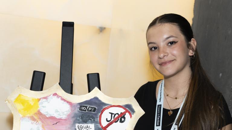 A young woman stands next to a shield painted with sunny skies and a boat floating on water on one side, and a dark sky and a sinking boat.