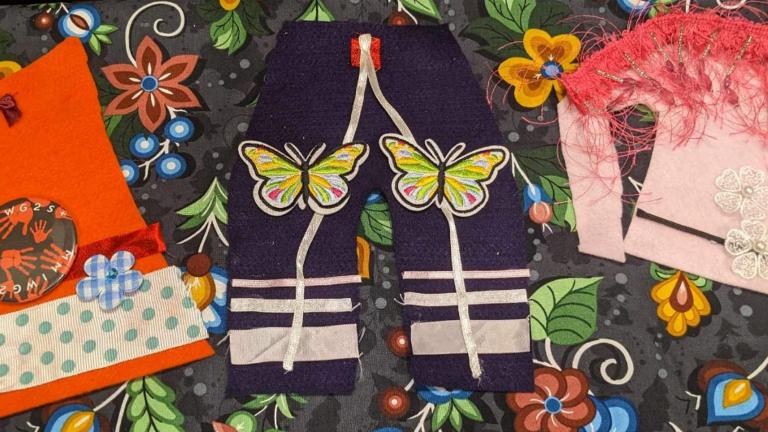 Pieces of crafted Indigenous- style clothing, including an orange shirt, pair of pants and made from colourful fabric and ribbon is are laid against a piece of craft paper adorned with flowers and berries. The clothing is includes an orange shirt, a pair of pants with ribbons and a pink tunic decorated with colourful fabric, appliqués and ribbons. The tunic has with a feathered shawl on the shoulders. Partially obscured.
