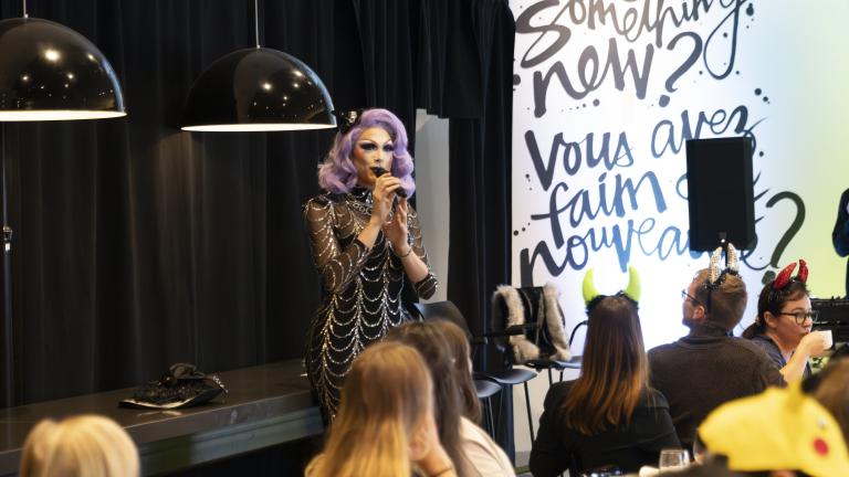 A drag performer with purple hair and a silver and black dress speaks in front of a group having brunch. Partially obscured.