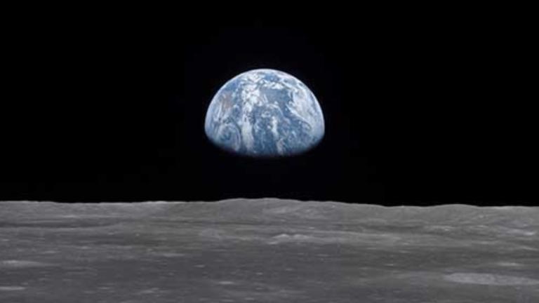 View of earth rising above the lunar horizon, showing the blue and white planet against the dark backdrop of space as seen from the moon’s surface. Partially obscured.