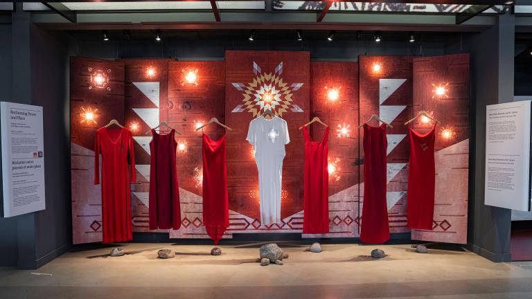 Six red dresses and a white dress in the middle are suspended in air on hangers in front of a backdrop. There’s a projected golden star placed on the heart of the white dress. The backdrop features red-painted birch wood panels with a white and yellow sun in the middle and white geometrical shapes on the last panels. Partially obscured.