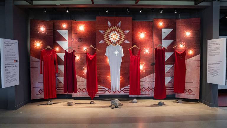 Six red dresses and a white dress in the middle are suspended on wooden clothes hangers in front of a backdrop. A white star glows at the location of the heart on the central white dress. The backdrop features patterns on a birchbark texture. Some of the patterns are illuminated from behind to create glowing star-like features on the panels. Partially obscured.