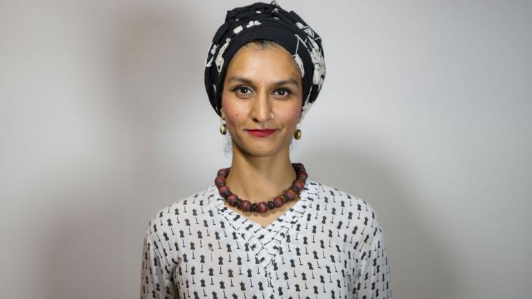 A Pakistani woman, Anam Abbas, in a black and white headwrap, red lipstick, chunky brown collar necklace and black-and-white blouse looks forward with a bemused expression. Partially obscured.