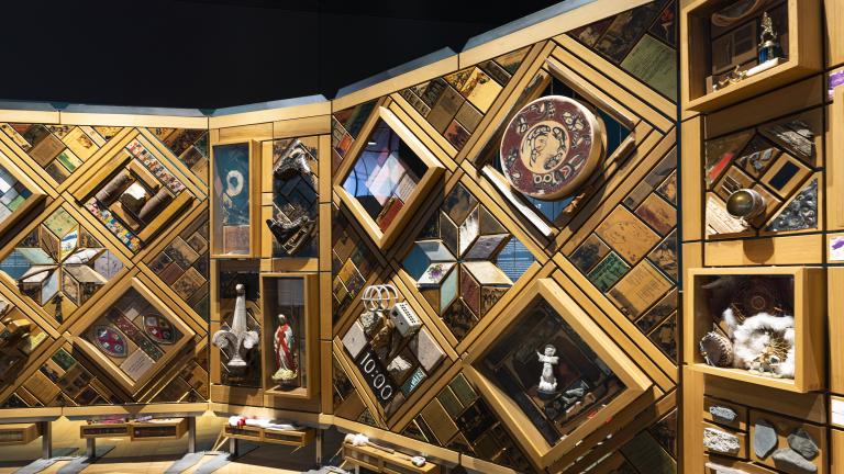 A large art installation composed of hundreds of objects embedded in a cedar frame resembling a quilted blanket. Partially obscured.