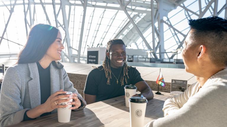 Three people sit around a table, smiling, each with a paper coffee cup in front of them. The sun shines behind them, through the panes of glass that wrap around the Canadian Museum for Human Rights. Partially obscured.