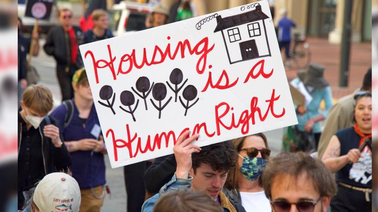 A group of people walking down a street. One person holds a sign that reads: “Housing is a human right.” Partially obscured.