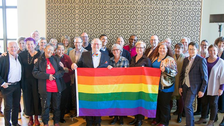 Twenty-eight members of the LGBT Purge Fund Board and LGBTQ2+ National Monument Advisory Committee standing together and holding a rainbow flag, at the first Monument visioning session in 2019. Partially obscured.