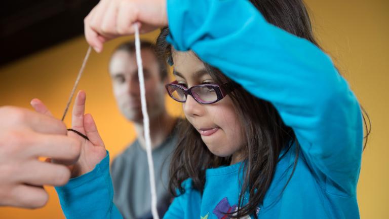 A child making a craft works intently to pull a string. The child is dressed in a blue sweatshirt with a snowman on it. Partially obscured.