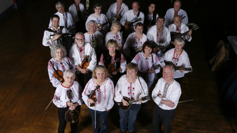 A group of twenty-two people facing the camera and smiling. They are all wearing dark-coloured pants and white shirts with red embroidery. A majority of the group are holding their instruments, mainly mandolins. Partially obscured.