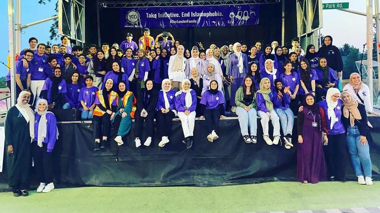 Dozens of people are gathered on a stage, most smiling and wearing the colour purple. Many are wearing hijabs. A banner hangs at the back of the stage that reads "Take Initiative. End Islamophobia. #OurLondonFamily." Partially obscured.