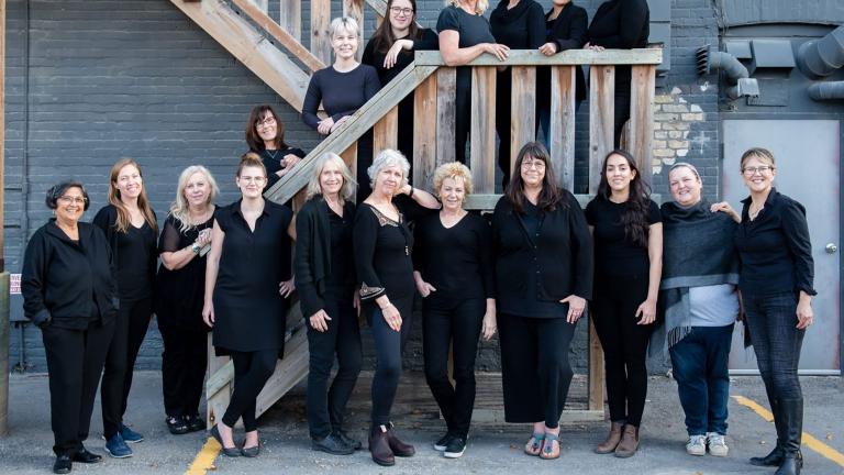 Women’s choir members are dressed in black and posed along an outdoor wooden staircase. Partially obscured.