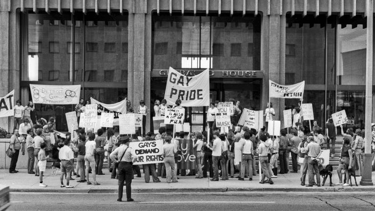 A group of people protesting on the sidewalk in front of an office building. They are holding protest signs and banners and are arranged three and four deep. A police officer watches them.