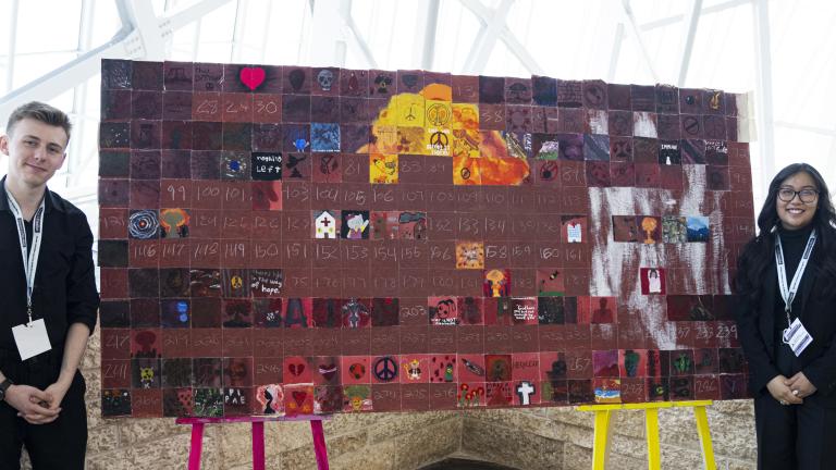 A young man and young woman stand in front of a mural made up of many squares. Some of the squares are filled with imagery, while others are filled with a brown background, waiting to be filled.