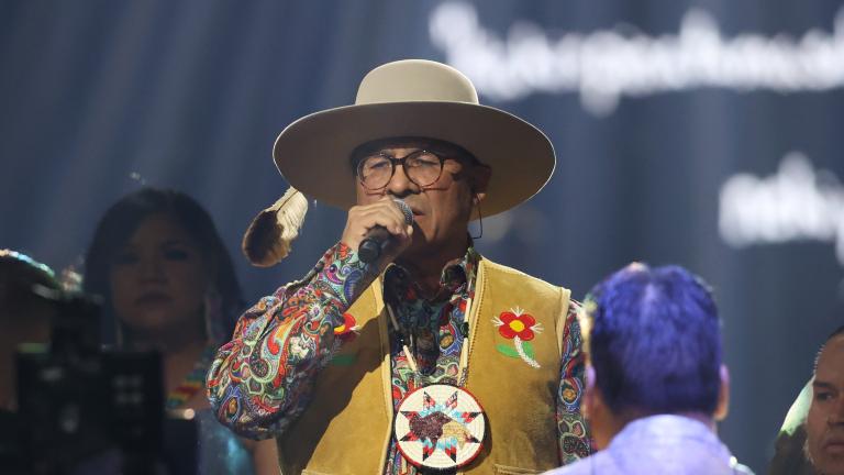 Close-up on a group of people wearing beaded jackets and vests performing on stage. In the centreer, a man wearing glasses, a hat, and a large, beaded medallion sings into a microphone. Partially obscured.