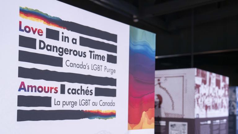 A large sign is made up of thick black horizontal lines and the English and French words “Love in a Dangerous Time: Canada’s LGBT Purge” and “Amours cachés : La purge LGBT au Canada” are distributed within the lines. The top and bottom lines are a mix of black and rainbow colours.