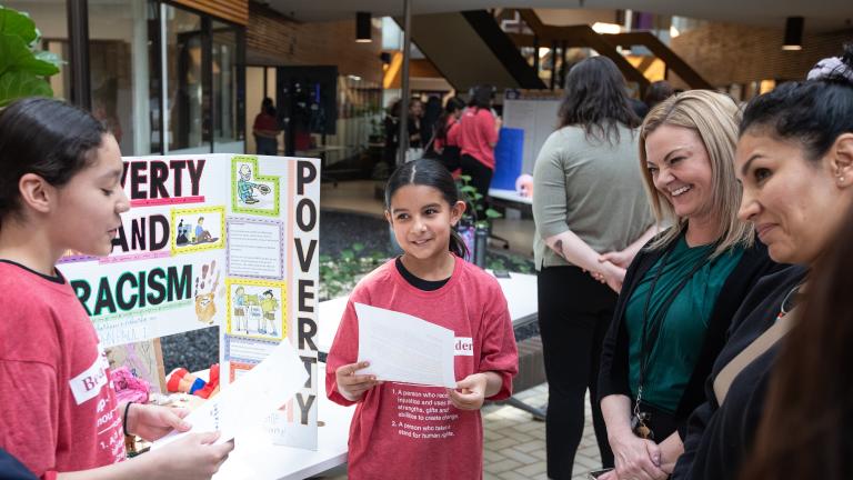 Two students standing in front of their project displayed on a table, presenting their work to visitors. Partially obscured.