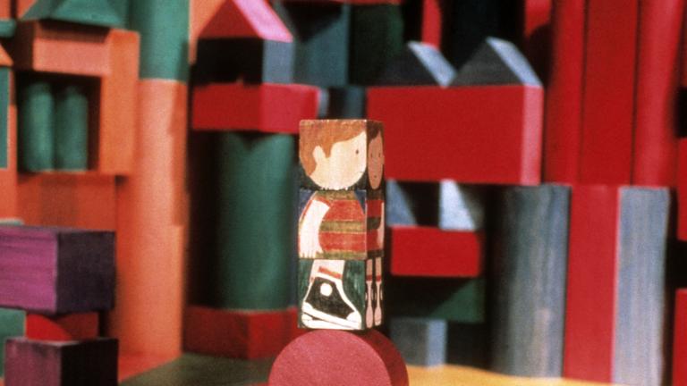 A wooden block painted with the image of a child, stands on top of a round block. Blocks of varying shapes, colours and sizes appear in the background.