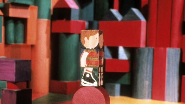 A wooden block painted with the image of a child, stands on top of a round block. Blocks of varying shapes, colours and sizes appear in the background. Partially obscured.