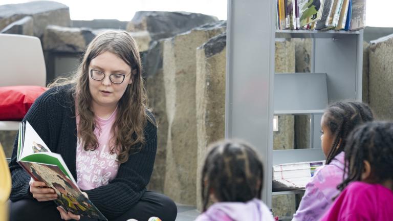 An adult sitting on the floor reading a book to three children sitting in front of them. Partially obscured.