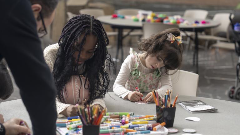 Two young children sit side by side at a round table, engaged in an art activity with markers and coloured pencils. Partially obscured.