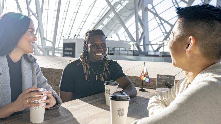 Three people are seated at a table. They're all smiling with coffees in front of them. There is a small progress pride flag on their table and at the empty table behind them. The sun is shining bright through the Museum windows. Partially obscured.