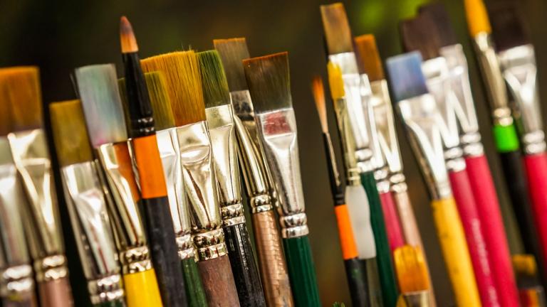 Various sizes of paint brushes standing in a paint-brush holder against a blurred green backdrop. Partially obscured.
