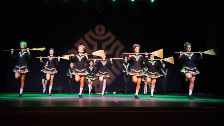Thirteen Irish dancers dressed in short, black dresses are holding brooms across their chest. Partially obscured.