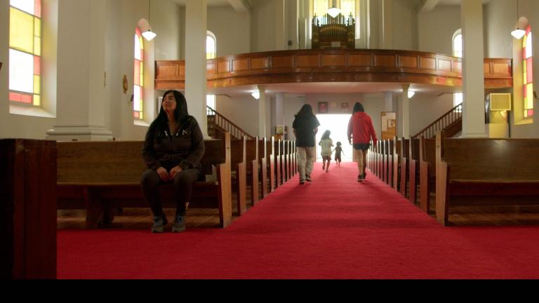 A church interior with a long red-carpeted aisle between wooden pews. Sunlight filters through stained-glass windows as several children run toward the bright entrance at the far end while an Indigenous woman sits alone in a pew to the left. Partially obscured.