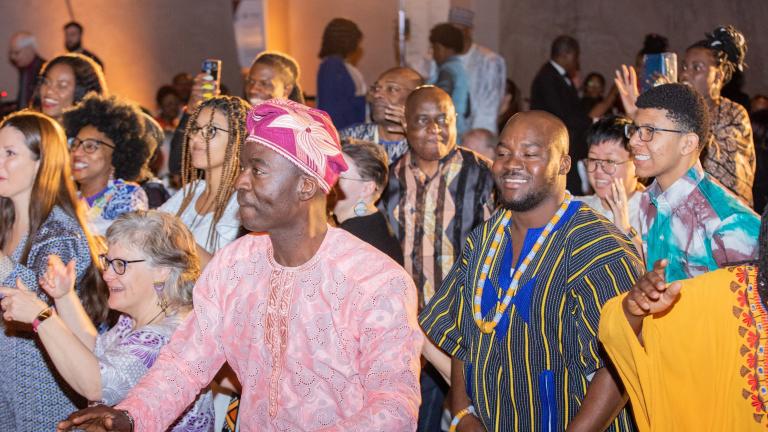 A group of people of all ages, many in brightly coloured traditional African attire, dancing and smiling. Partially obscured.