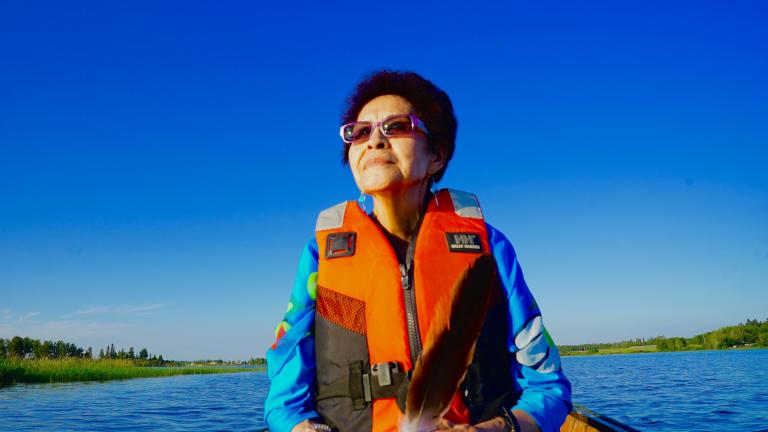An indigenous woman is in a canoe on a lake with an orange life jacket. She seems at serene. Partially obscured.