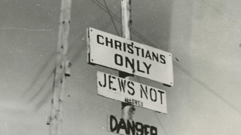 Black and white photo of three signs on a post, reading “Christians only,” “Jews not allowed” and “Danger.” Partially obscured.