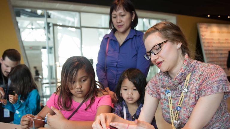 A woman strings yarn around her fingers as she demonstrates and explains what she is doing to two girls. Partially obscured.