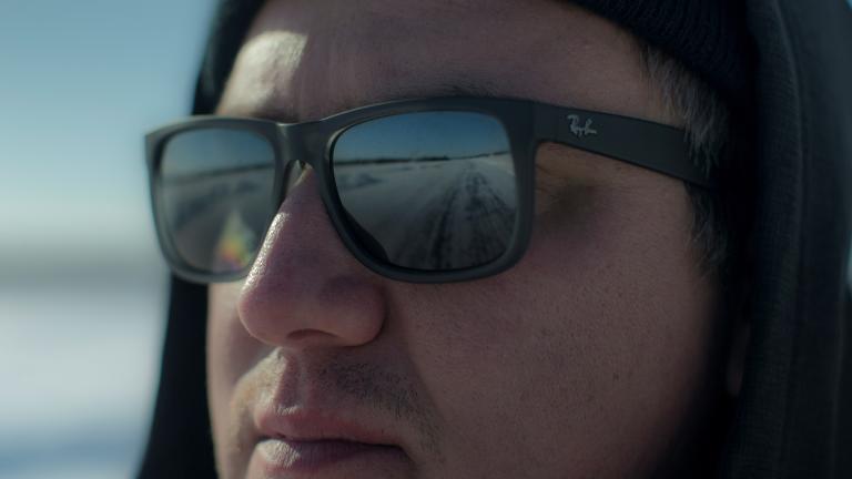 A close-up of a man wearing sunglasses and a hat, looking at something off camera. A road is seen reflected in his sunglasses. Partially obscured.