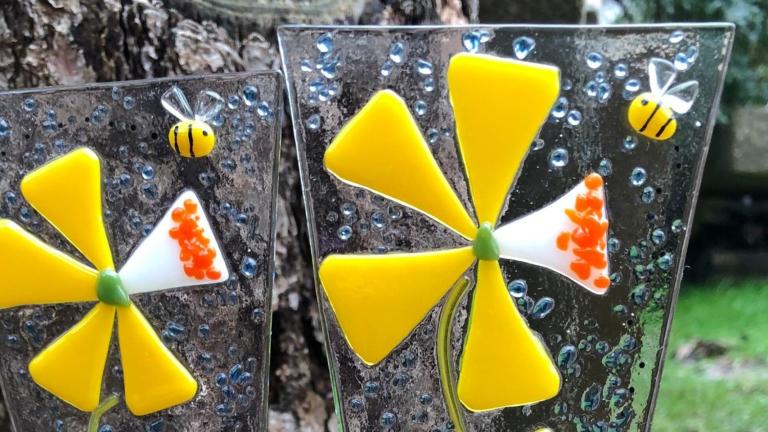 Two fused glass ornaments depicting yellow flowers sit in a flower pot outside. Partially obscured.