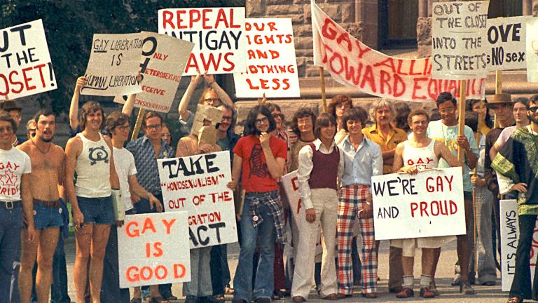 A large group of people dressed in T-shirts and seventies-style flared pants, stand together holding signs with messages such as “Out of the Closet,” “Gay is Good” and “Repeal Anti-Gay Laws.” Partially obscured.
