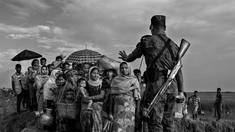 A soldier with a rifle on his back blocks a group of Rohingya before they cross a makeshift bridge. His left hand is raised, gesturing for them to stop. Partially obscured.