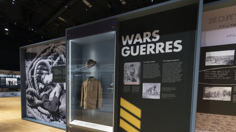 A series of exhibition panels feature historical images of military personnel. The word “Wars” is on the top of one panel. A display case located between two panels contains a First World War military uniform.