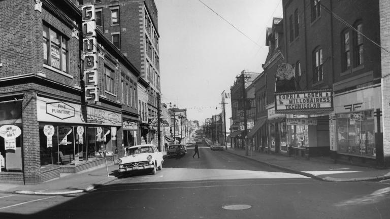 A black and white image of a town street. The street leads away from the viewer towards a vanishing point on the horizon, and is lined with brick buildings ranging from two to seven stories storeys tall. Many of the buildings have storefronts on the first floor, and the first building on the right has a movie marquee advertising a film called “The Millionairess” starring Sophia Loren.