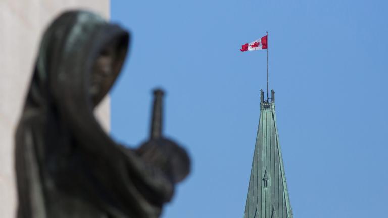 A Canadian flag flies atop a large tower. In the foreground stands a statue of a veiled woman carrying a sword. Partially obscured.