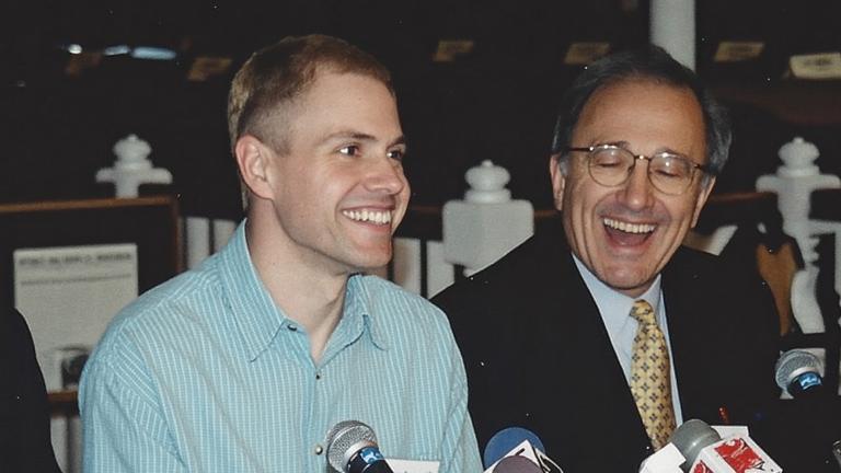 A man wearing a light blue shirt sits at a long table in front of a cluster of microphones, smiling. Seated on both sides of him are other people dressed in suits and facing microphones, also smiling. The name placards along the front of the table read, from left to right, “Murray Billet”, “Robinson Koilpillai”, and “Dr George Rodgers”, with too-blurry-to-read affiliations below the names. Another placard, knocked on its side, reads “Delwin Vriend” without an affiliation. Partially obscured.