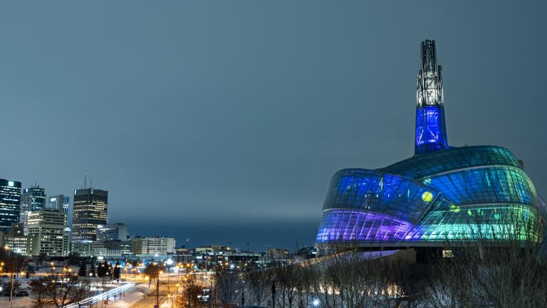 The CMHR’s exterior is seen at night with its glass panels lit up blue, green and purple. Partially obscured.
