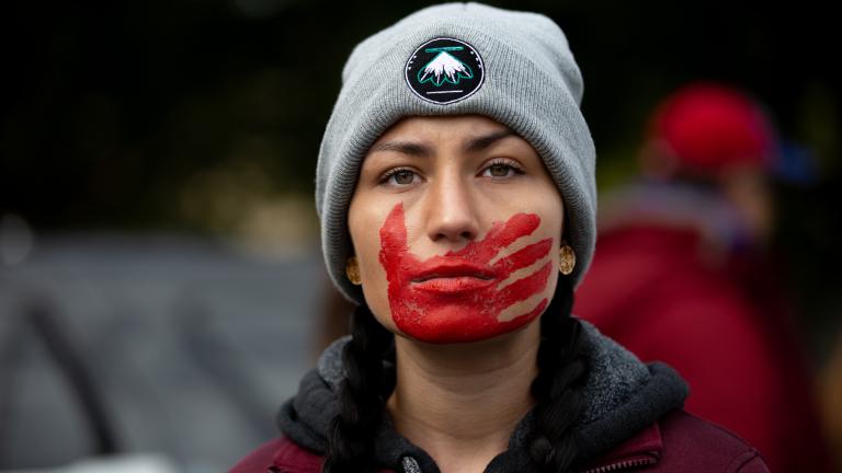 A young woman with braided hair, wearing a maroon jacket and gray winter cap, looks forward resolutely. A red handprint is painted over her mouth and face. Partially obscured.