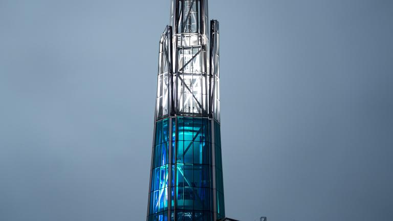 A tall, illuminated glass-and-steel spire rises above the curved patterned roof of the Canadian Museum for Human Rights, glowing in blue and white light against a soft grey sky. Partially obscured.