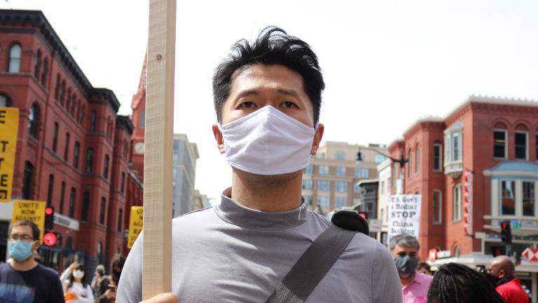 A man wearing a cloth mask and holding a sign, walking with a group of protestors carrying signs about ending racist violence and anti-Asian racism. Partially obscured.