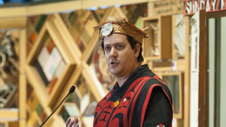 A man wearing traditional Indigenous regalia and headdress stands in front of an art installation made of wooden panels with embedded objects. Partially obscured.